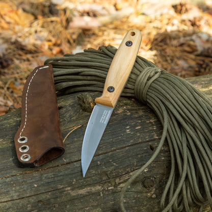 A fixed-blade knife with a light wood handle and silver blade rests on a log outdoors, next to a brown leather sheath and a coiled bundle of green cord. The background shows dry leaves and forest floor debris.