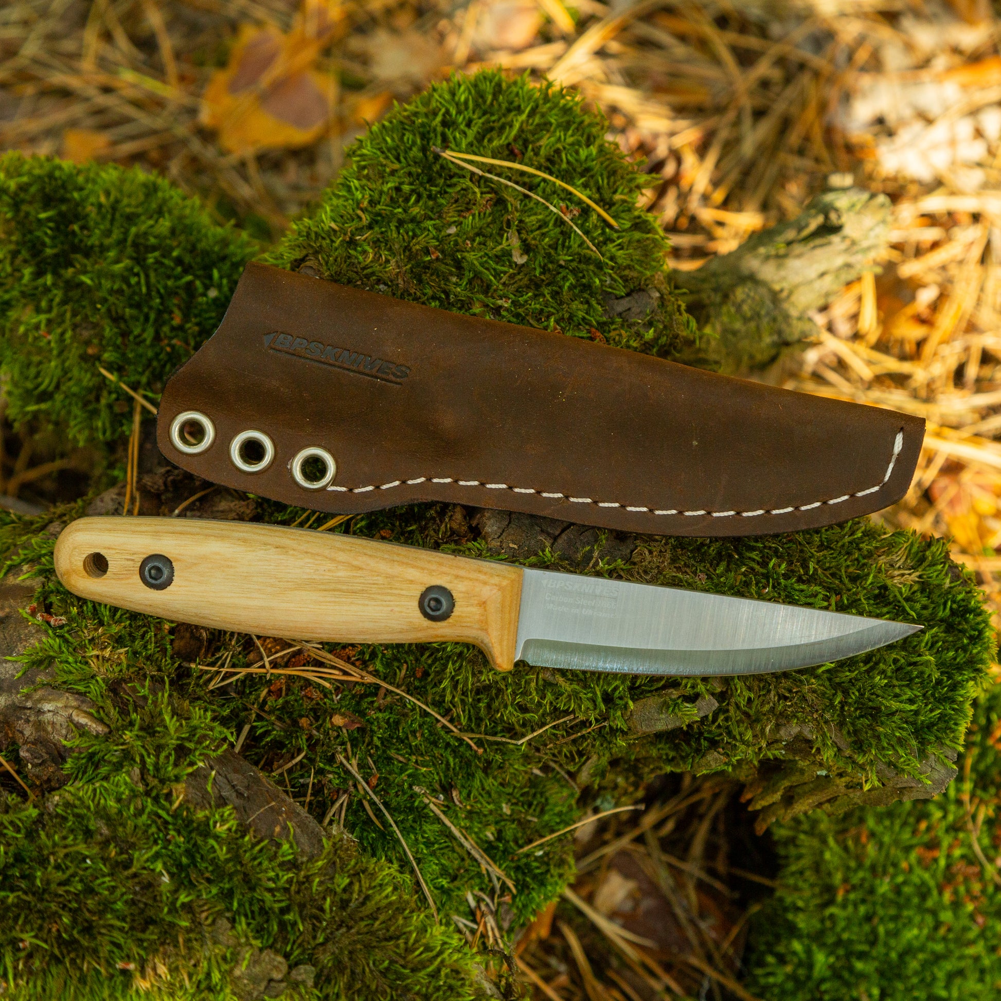 A fixed-blade knife with a light wood handle and silver blade lies on mossy logs outdoors, with its brown leather sheath placed above it. The sheath has visible white stitching and a debossed brand mark that appears to read 'Kings of NY'.