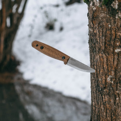 A fixed-blade knife with a brown wooden handle and a silver steel blade is shown embedded in a tree trunk outdoors, with snow and blurred forest in the background.